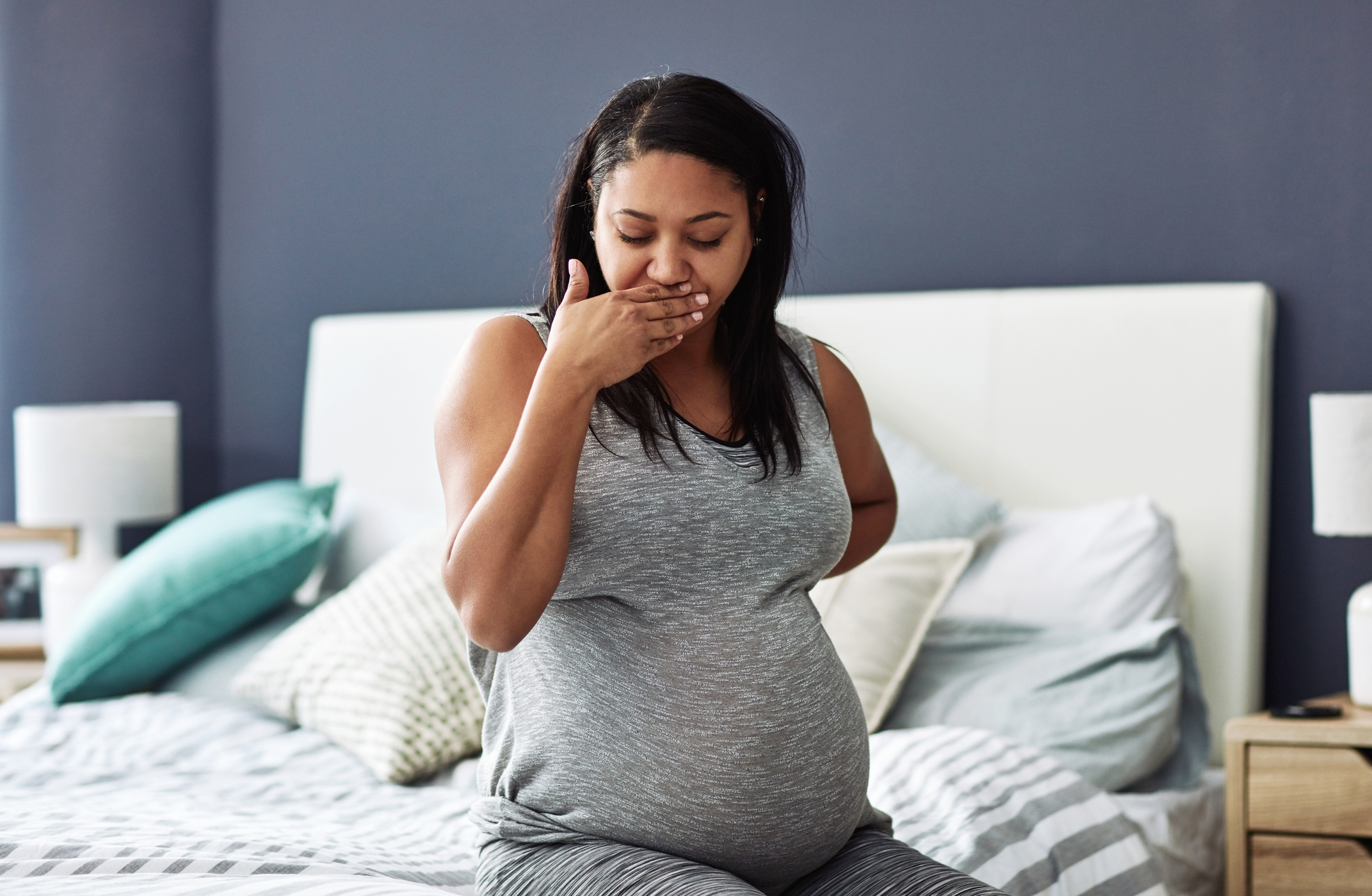A pregnant woman sits on the edge of her bed feeling sick