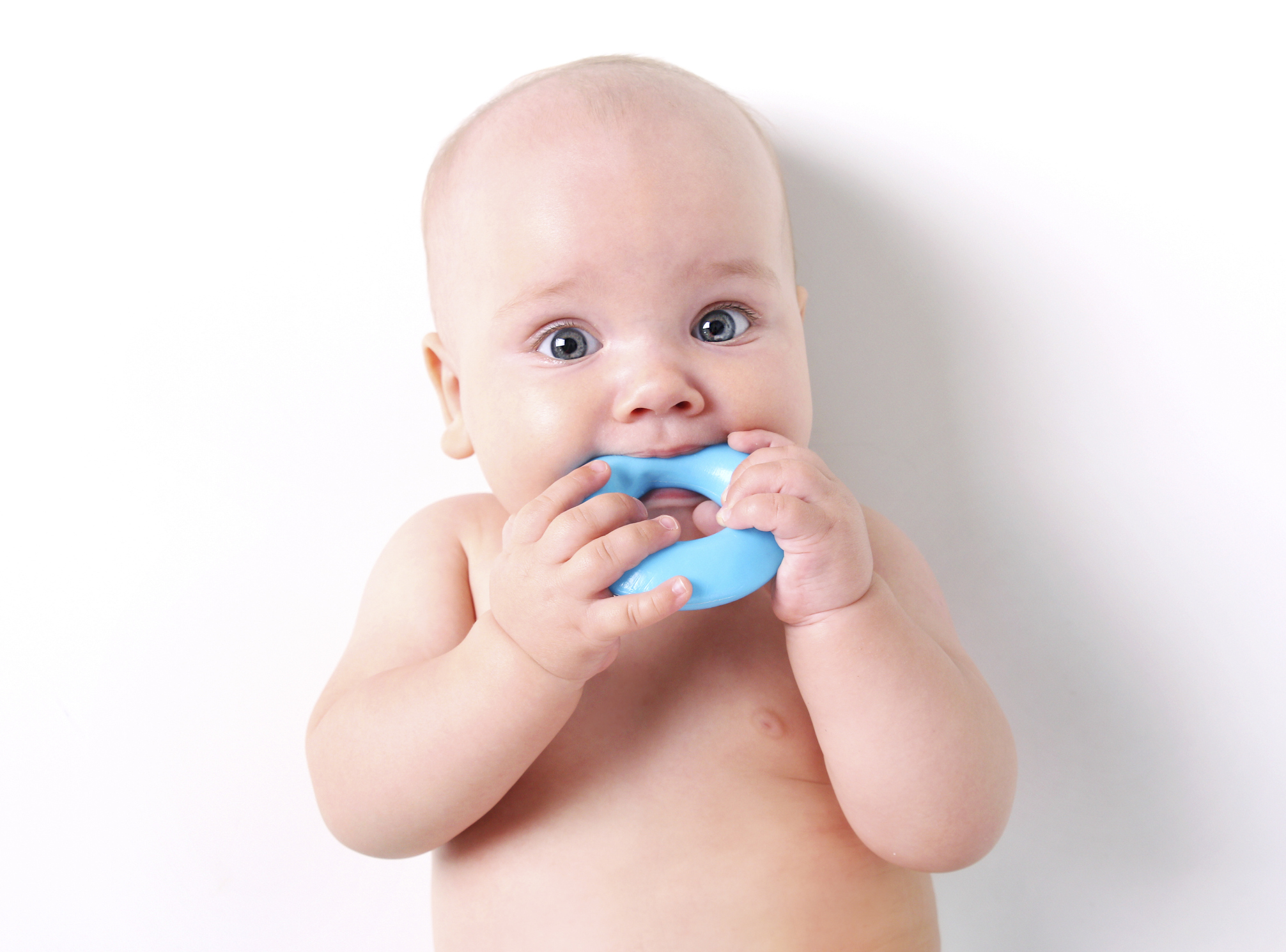 A baby lays on a white sheet chewing on a teething ring
