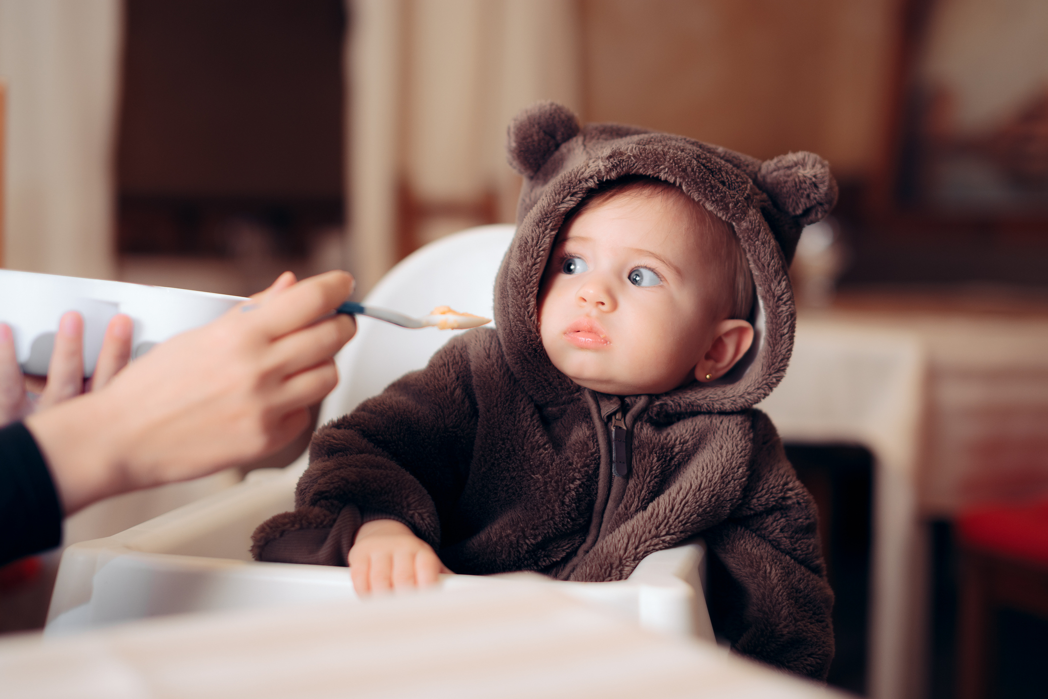 A baby in a bear onesie leans away from the spoon