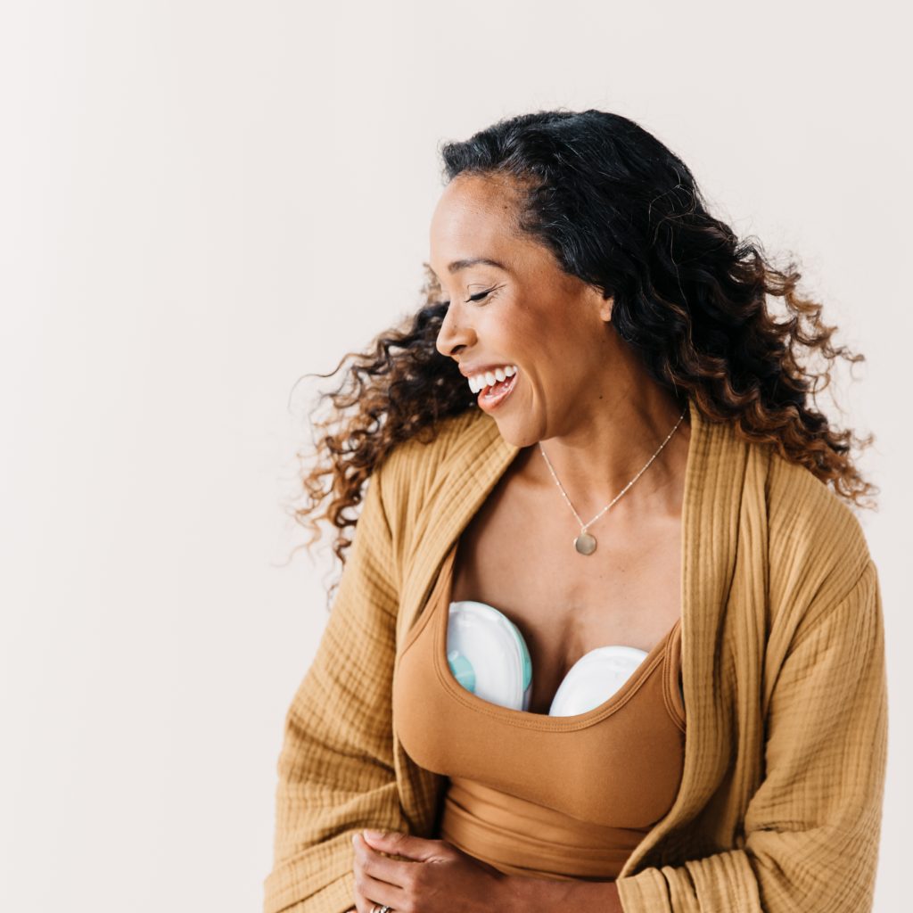 Woman with long hair smiles and looks to the side as she is wearing a tank top and cardigan with a hands-free breast pump tucked inside