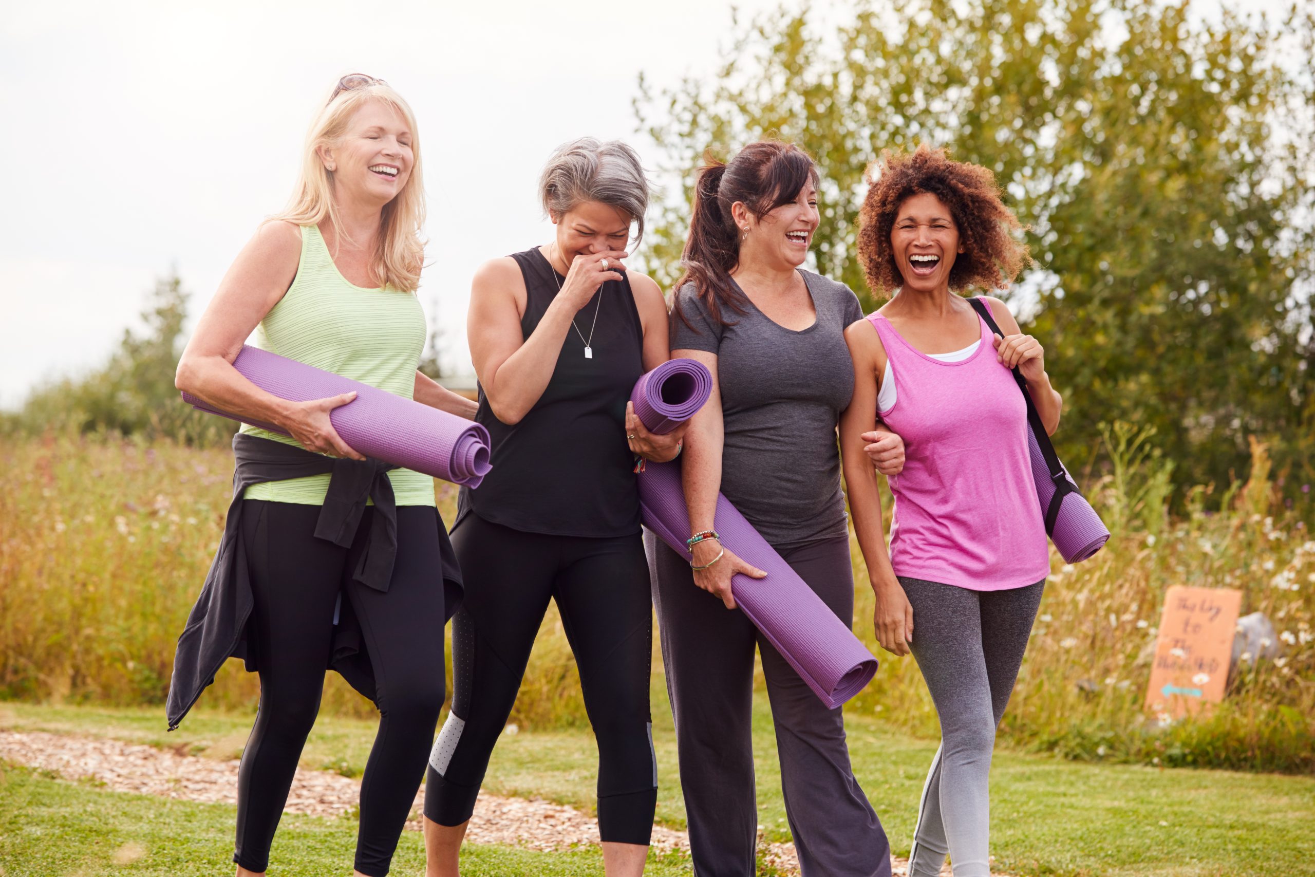 Group of four women walking outside holding yoga mats, laughing together