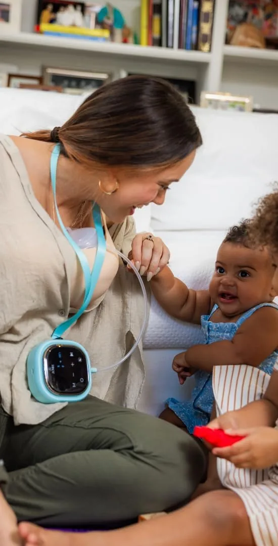 Mom wearing the Zomee Z2 on its lanyard with hands-free collection cups in her bra while she plays with her baby